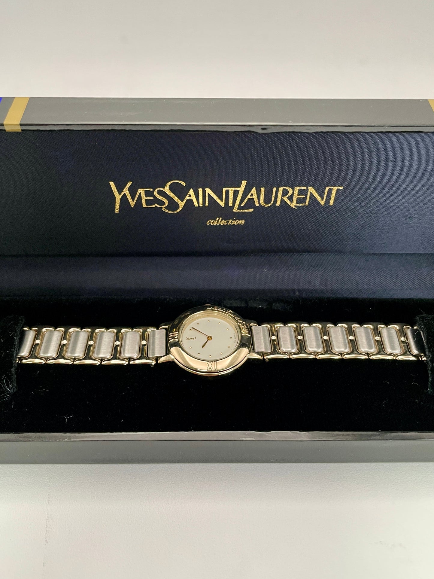 Horizontal view of YSL Pour Femme watch with gold case, black dial, and two-tone bracelet, shown with original box.