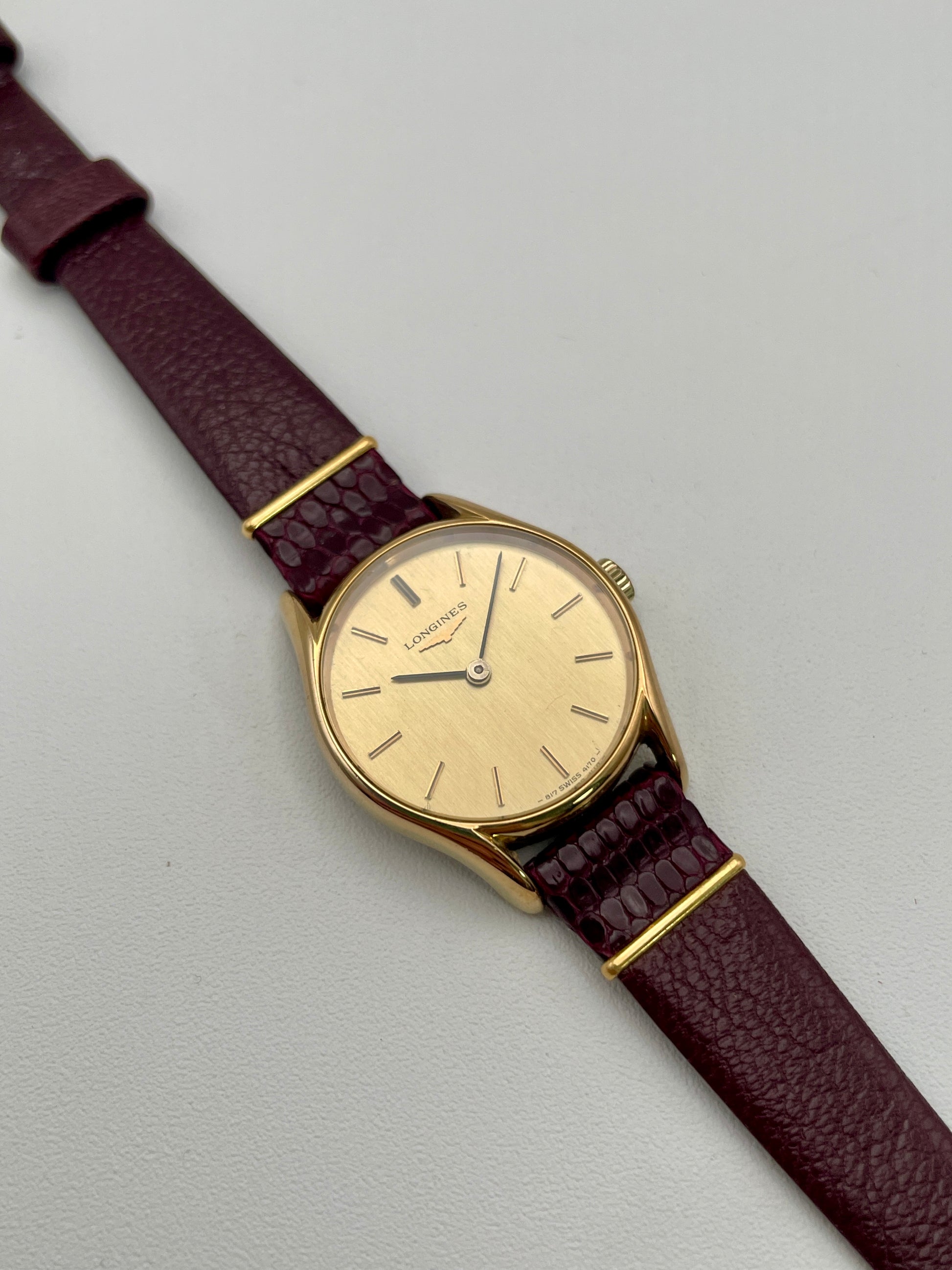 Longines Ref. 4139 vintage women's watch in gold with a burgundy leather strap, displayed diagonally to highlight its sunburst champagne dial.