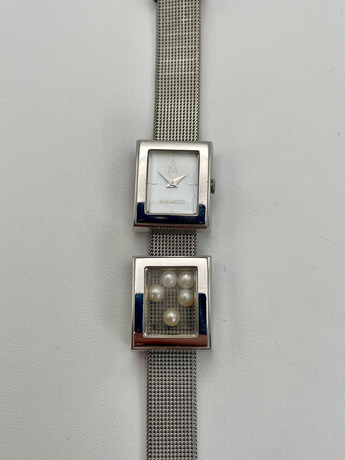 Front view of a Mikimoto ladies' watch featuring a rectangular silver-tone case, mother-of-pearl dial, and a unique design with a window on the bracelet containing loose pearls.