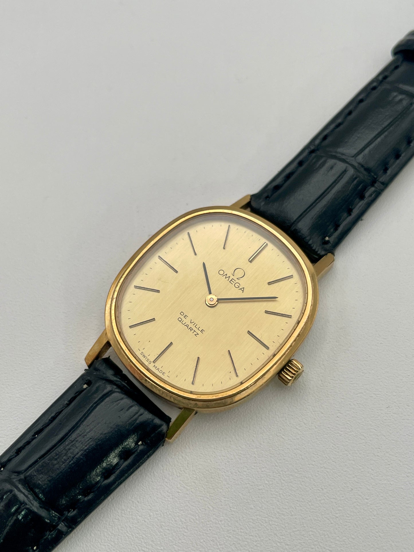 Diagonal view of the Omega De Ville Oval watch in gold with a black leather strap.