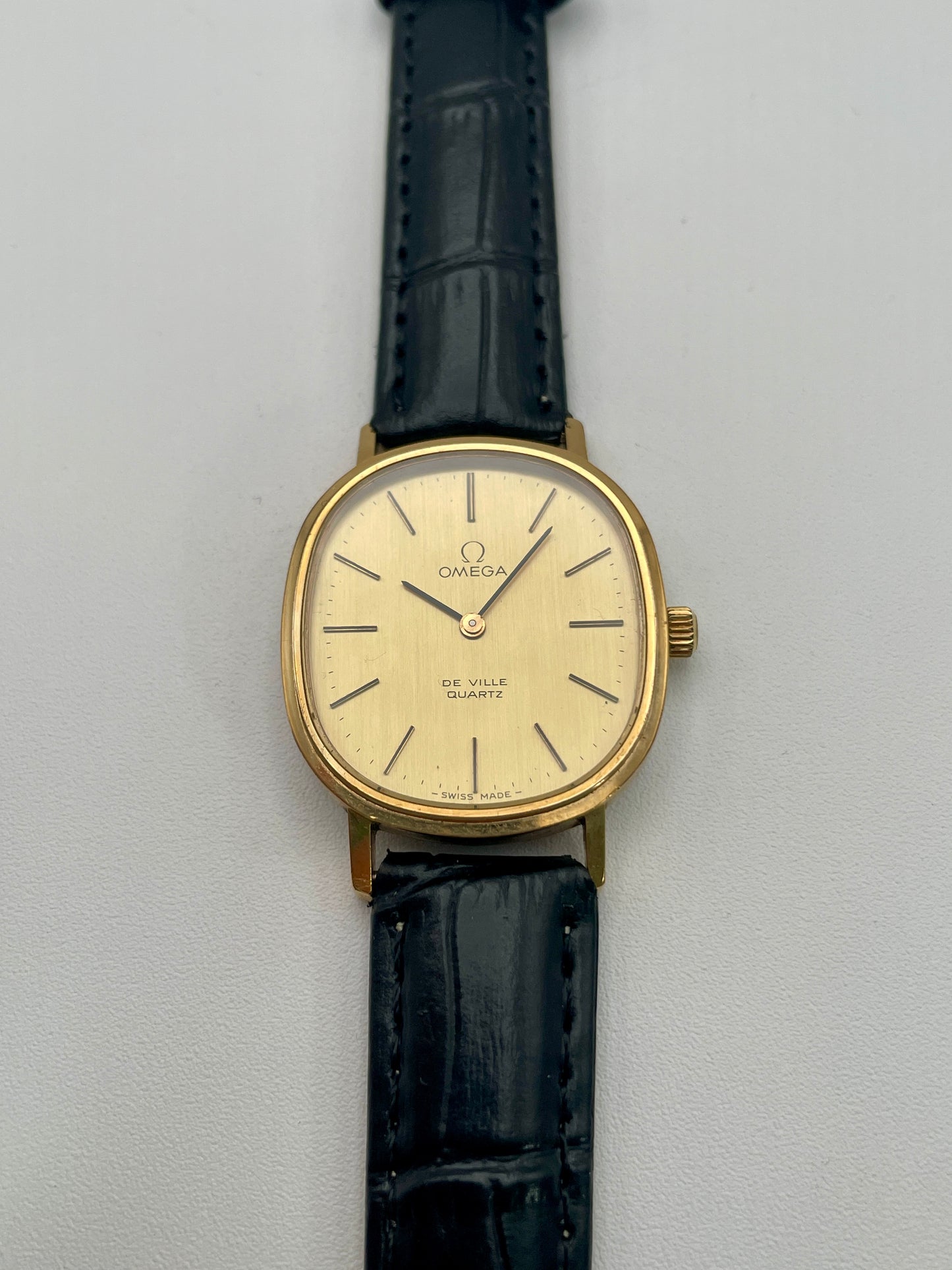 Omega De Ville Oval gold-tone watch showcasing its minimalistic dial and black leather strap, front view.
