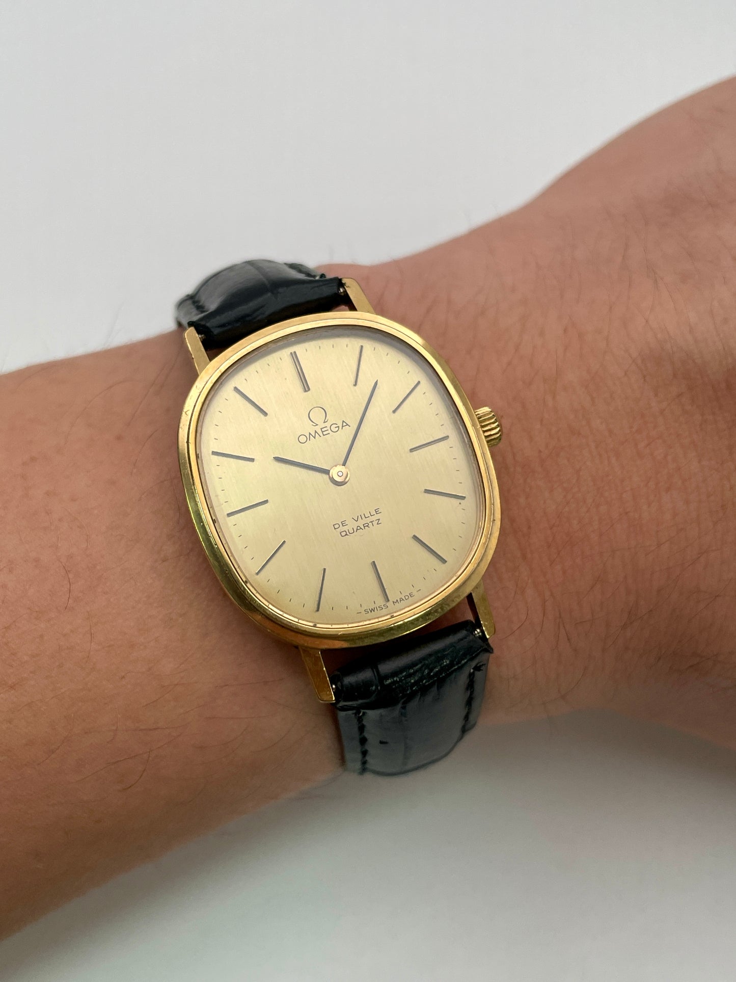 Omega De Ville Oval watch worn on the wrist, highlighting the gold case and black leather strap.
