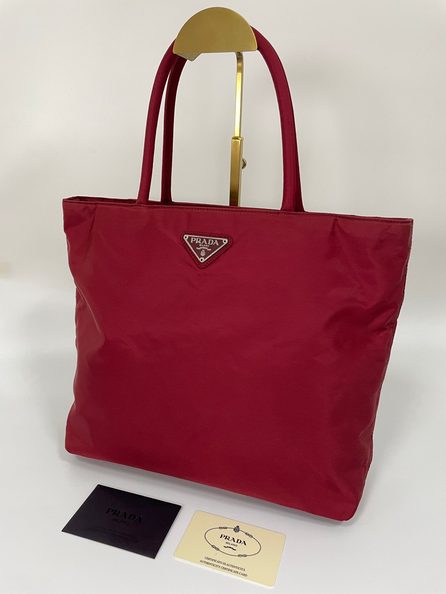 Angled view of a Prada Tessuto Tote Bag in red nylon with dual handles and the Prada triangle logo, displayed with authenticity cards.