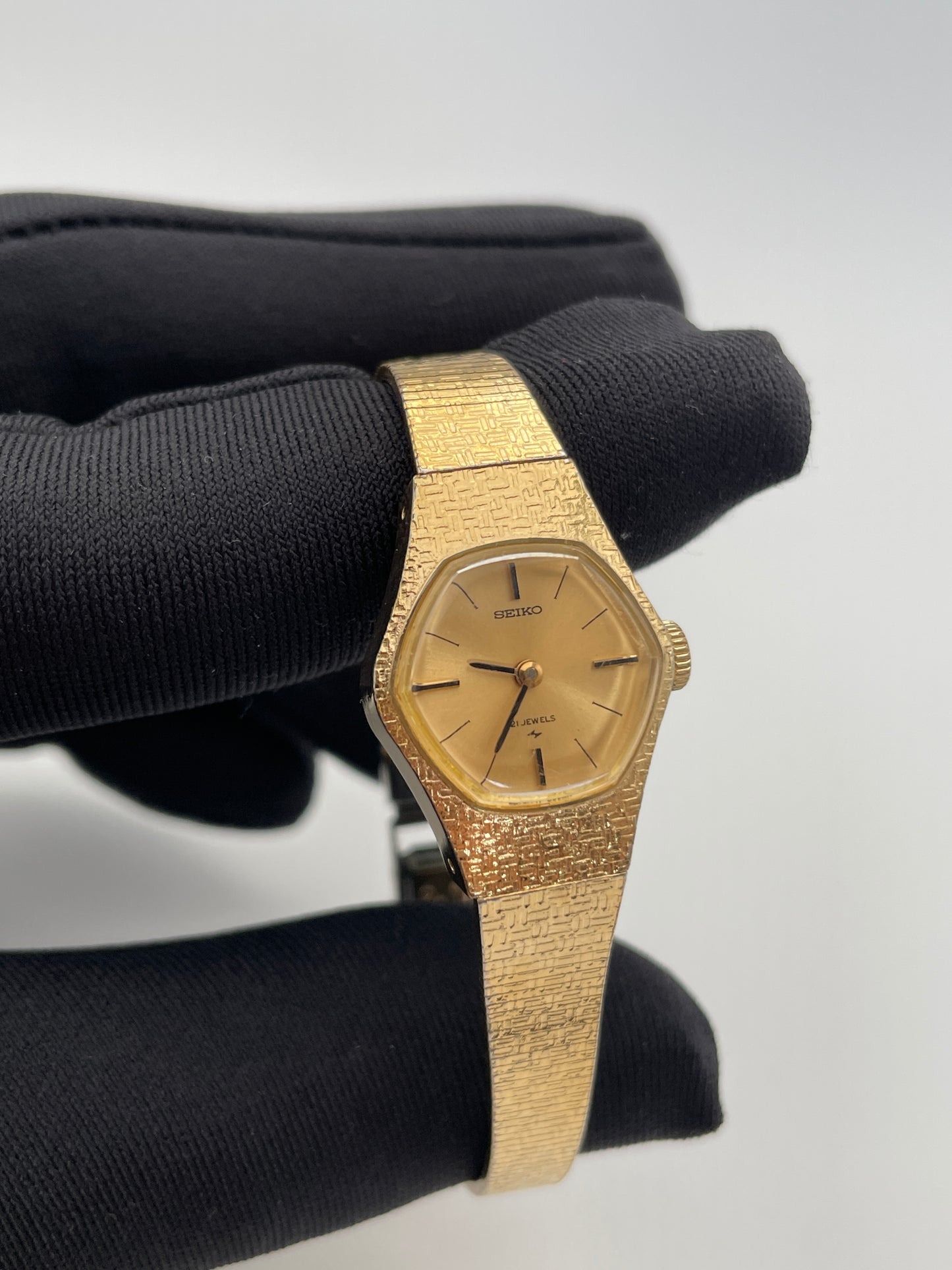 ront view of a vintage Seiko ladies' watch held in a gloved hand, featuring a gold-tone textured case and bracelet with a simple gold dial, black hour markers, and hands.
