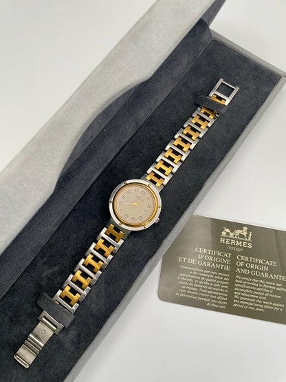 Tiew of a Hermes Clipper 30mm two-tone watch with a stainless steel and gold bracelet in its original Hermes box and guarantee. The silver dial features Arabic numerals and a date window at the 3 o'clock position.