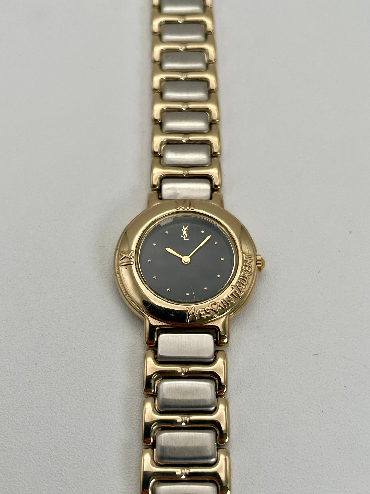 Front view of YSL Pour Femme watch with gold case, black dial, and two-tone bracelet.