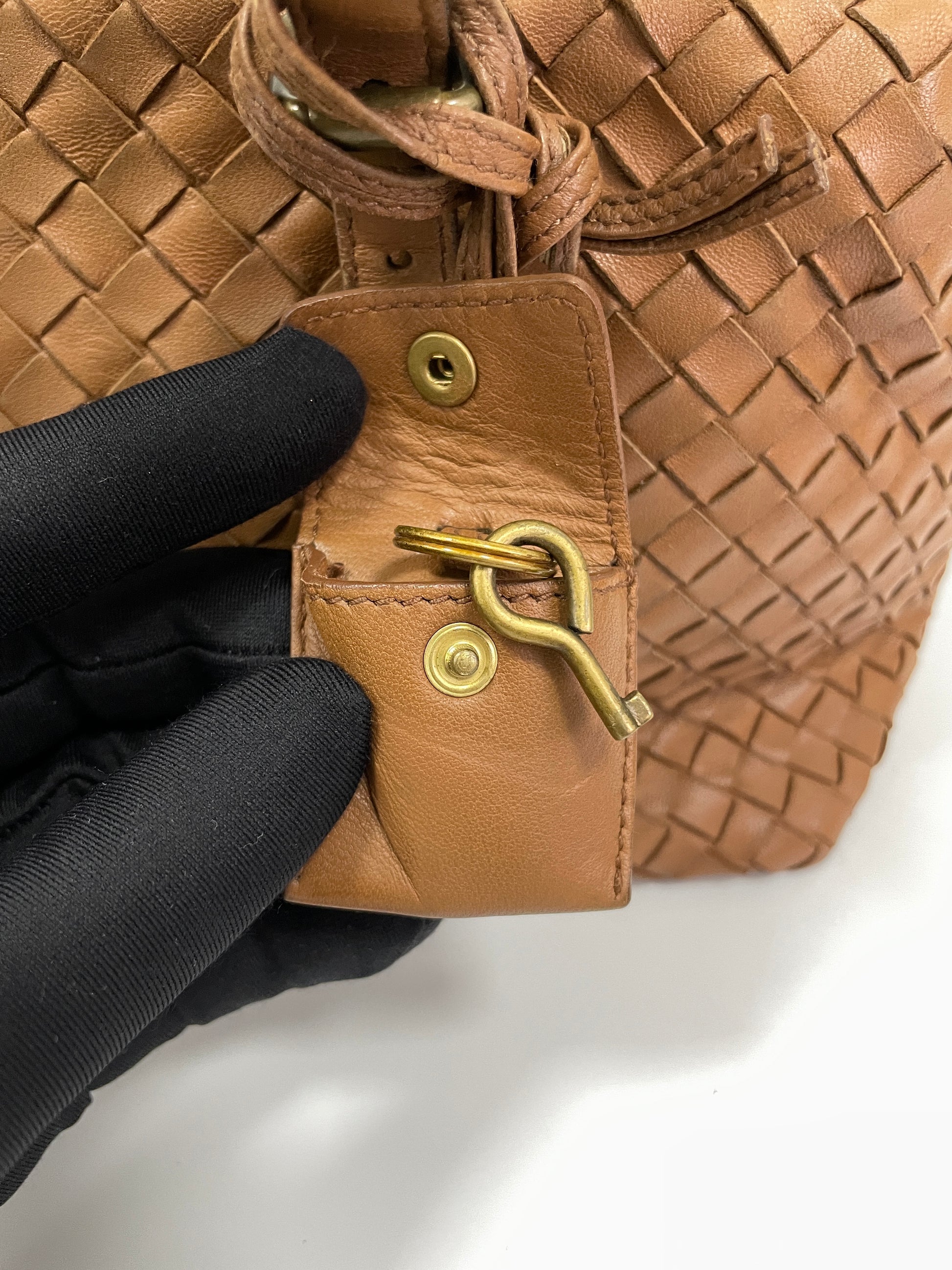 Close-up view of the brass clochette key detail on the Bottega Veneta Intrecciato Montaigne bag in brown leather.

