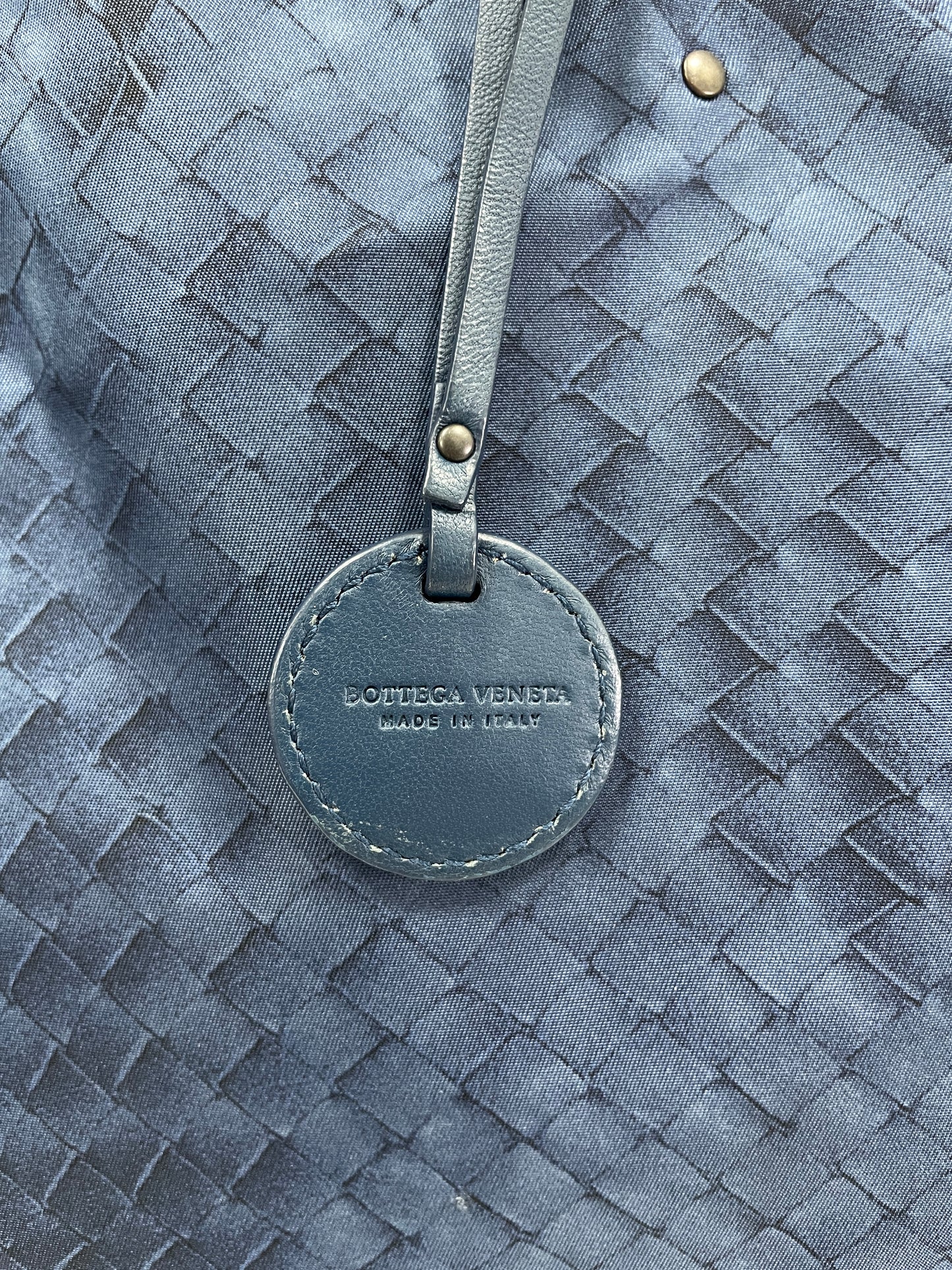 Close-up of the Bottega Veneta logo on the leather tag of the Bottega Veneta Intrecciolusion Mirage Butterfly Tote Bag in blue, stamped with "Made in Italy."