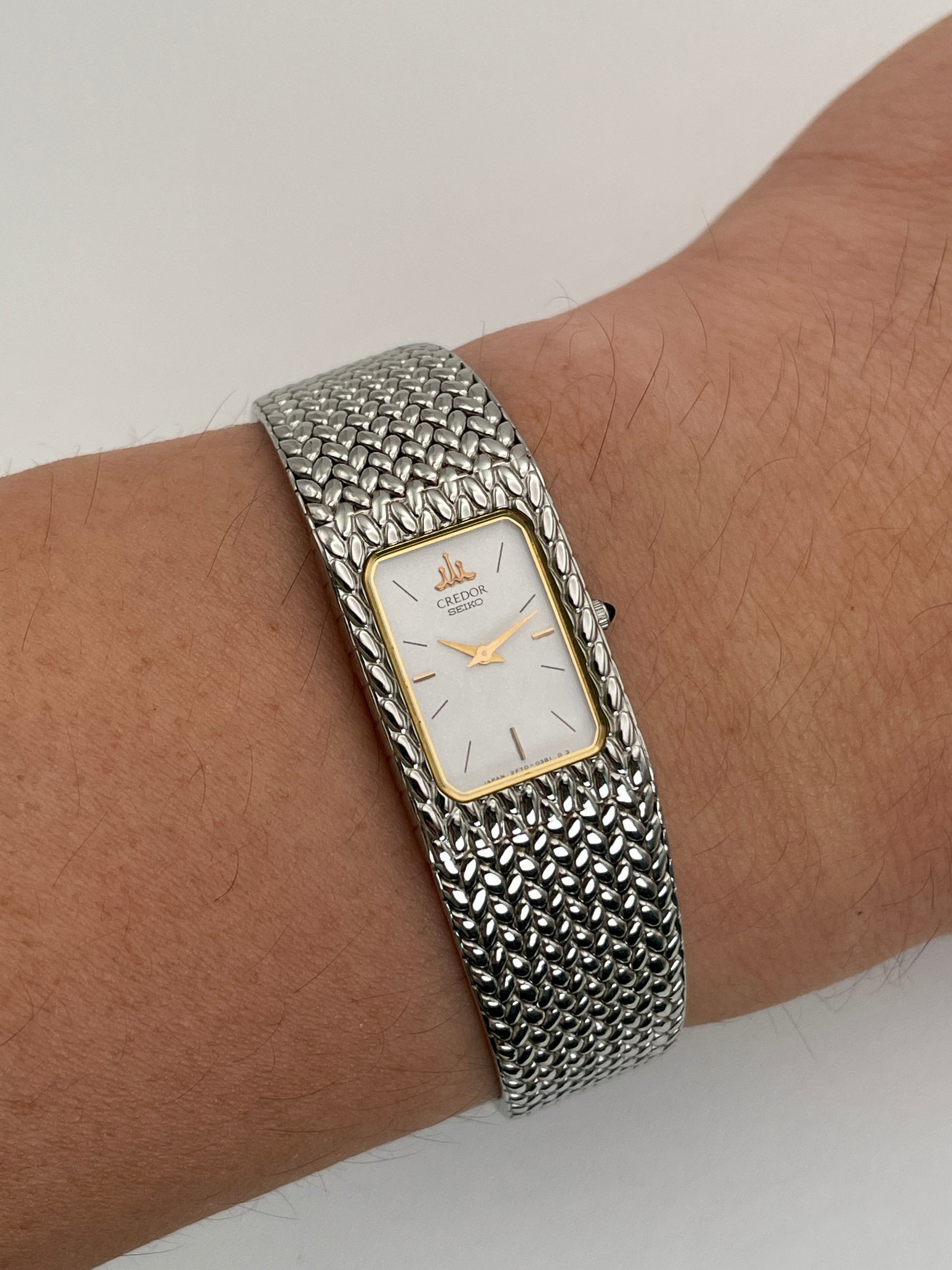 Wrist shot view of a Credor 2F70 bracelet watch with a silver-tone rectangular case, gold-tone bezel, white dial, and a textured mesh bracelet, elegantly worn on the wrist.