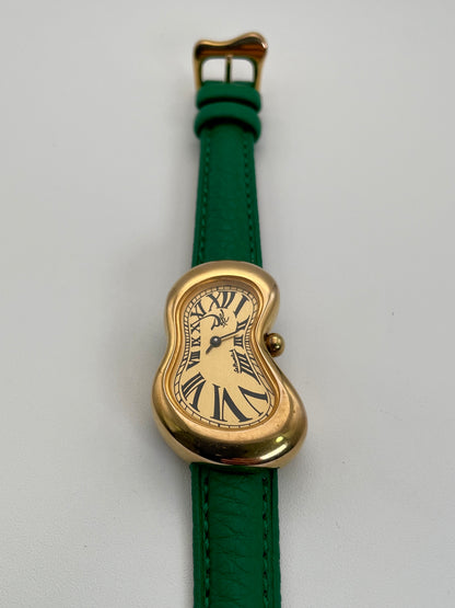 Front view of an Exaequo Salvador Dali Softwatch with a gold-toned, distorted case, Roman numeral dial, and green leather strap.