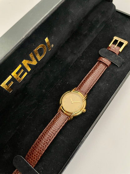 Top view of Fendi 300L watch with gold-tone case and brown leather strap, displayed in a black Fendi box.