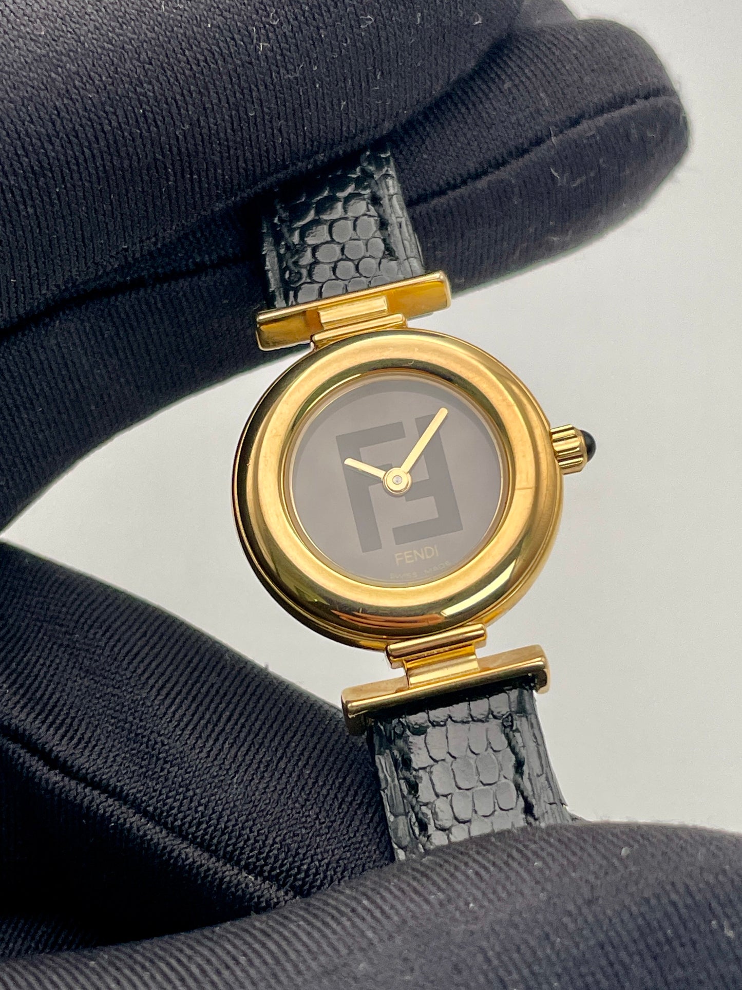 Close-up view of the Fendi 320L watch with a gold-tone round case, black dial with Fendi logo, and black leather strap