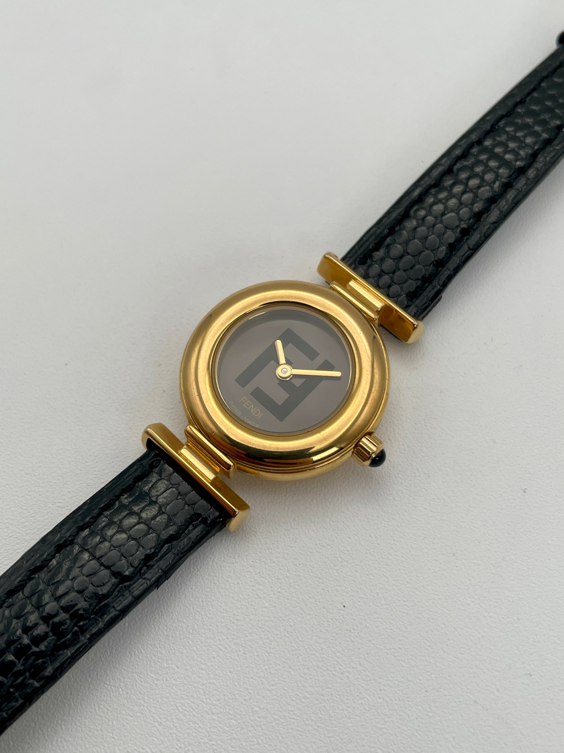 Diagonal view of the Fendi 320L watch highlighting the gold-tone round case, black leather strap, and black dial