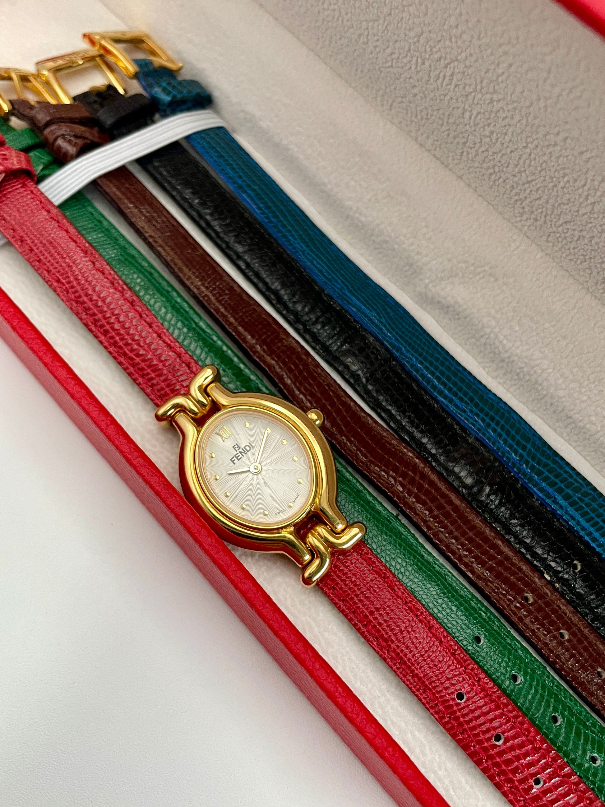 Fendi 640L gold watch displayed in a box with five interchangeable leather straps, including red, green, and black.