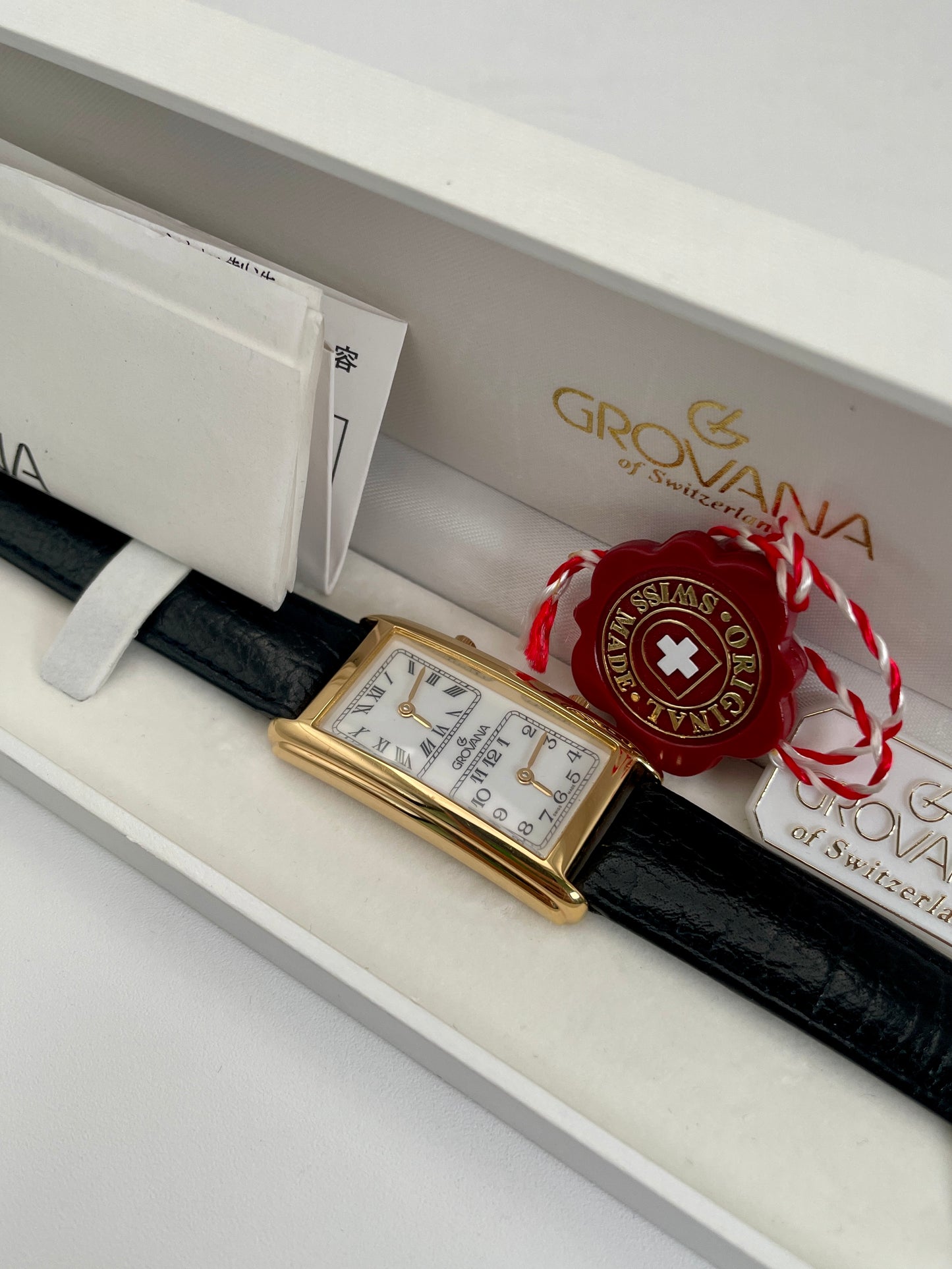 Grovana Tank Dual Time watch with a gold-tone rectangular case, white dual dials, and black leather strap, displayed in the original Grovana box.