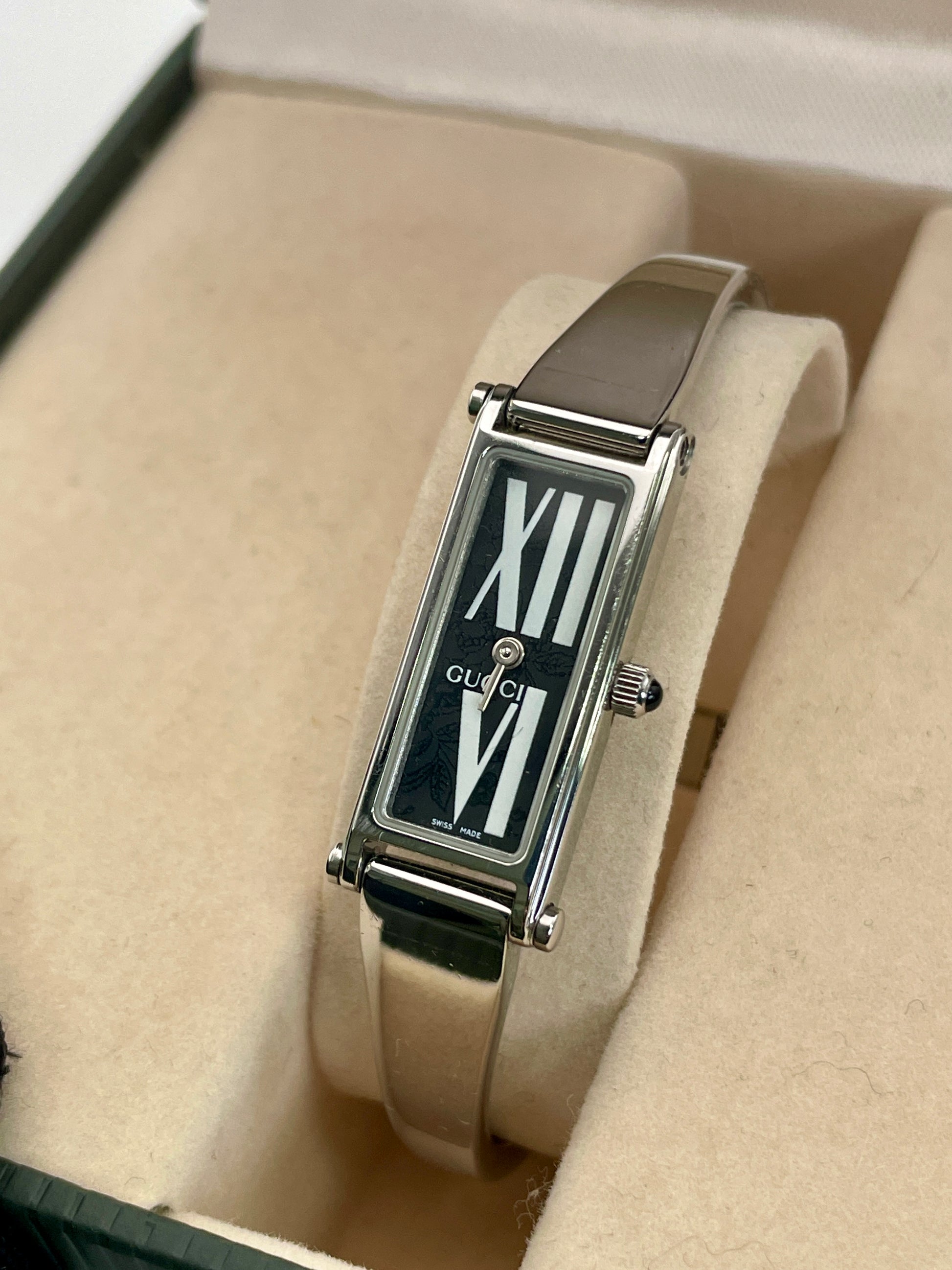 Close up view of Gucci 1500L black dial watch with a rectangular silver-tone case and stainless steel bracelet, displayed in the original Gucci box.