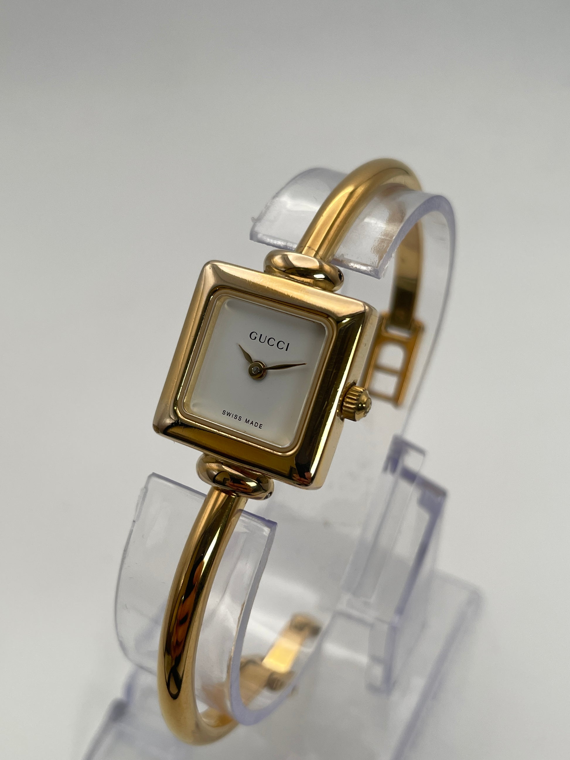 Diagonal view of Gucci 1900L gold watch with a square case and white dial, featuring a gold bangle bracelet.