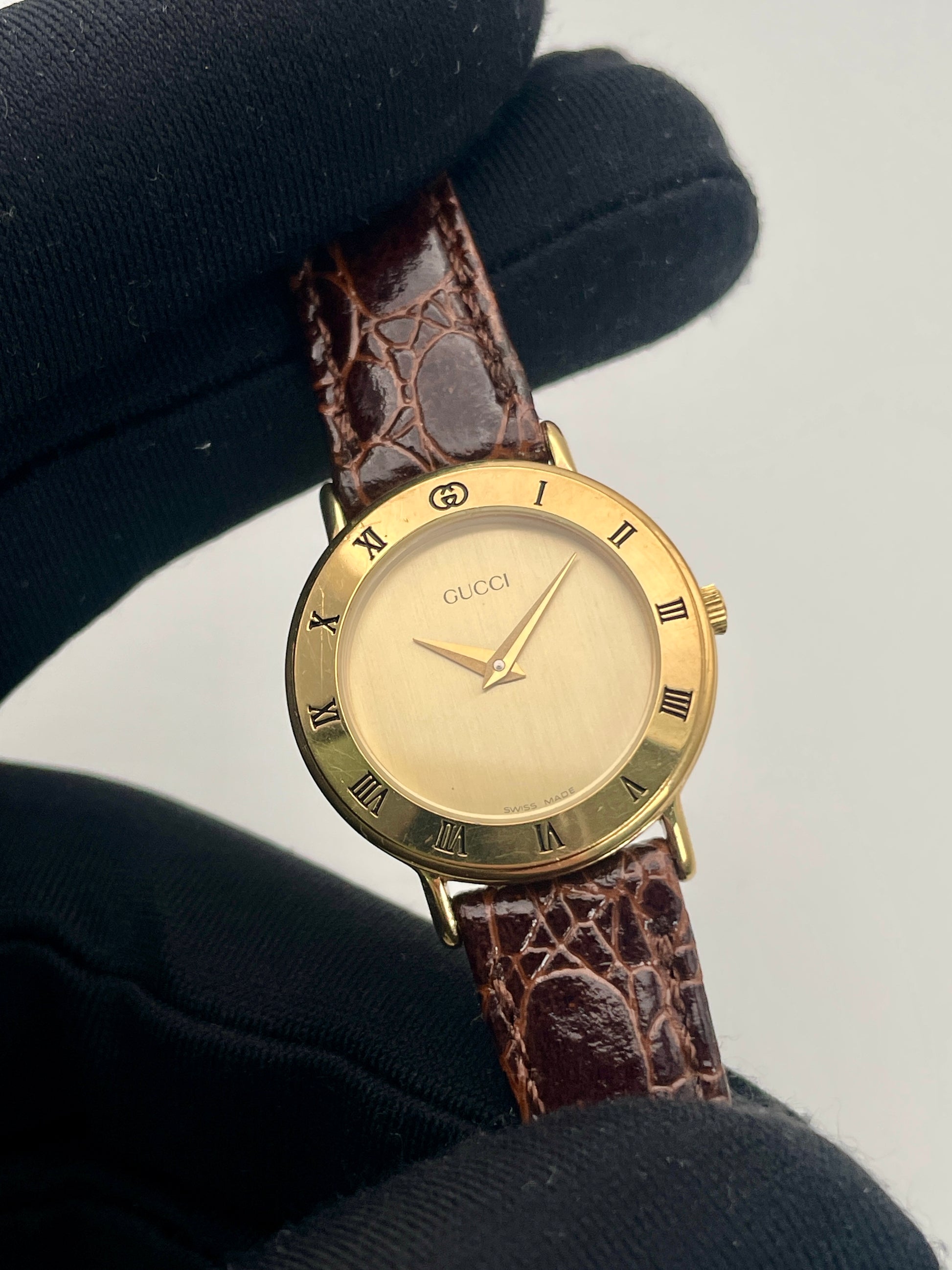 Close-up view of the Gucci 3000.2.L watch with a gold-tone bezel and brown leather strap.

