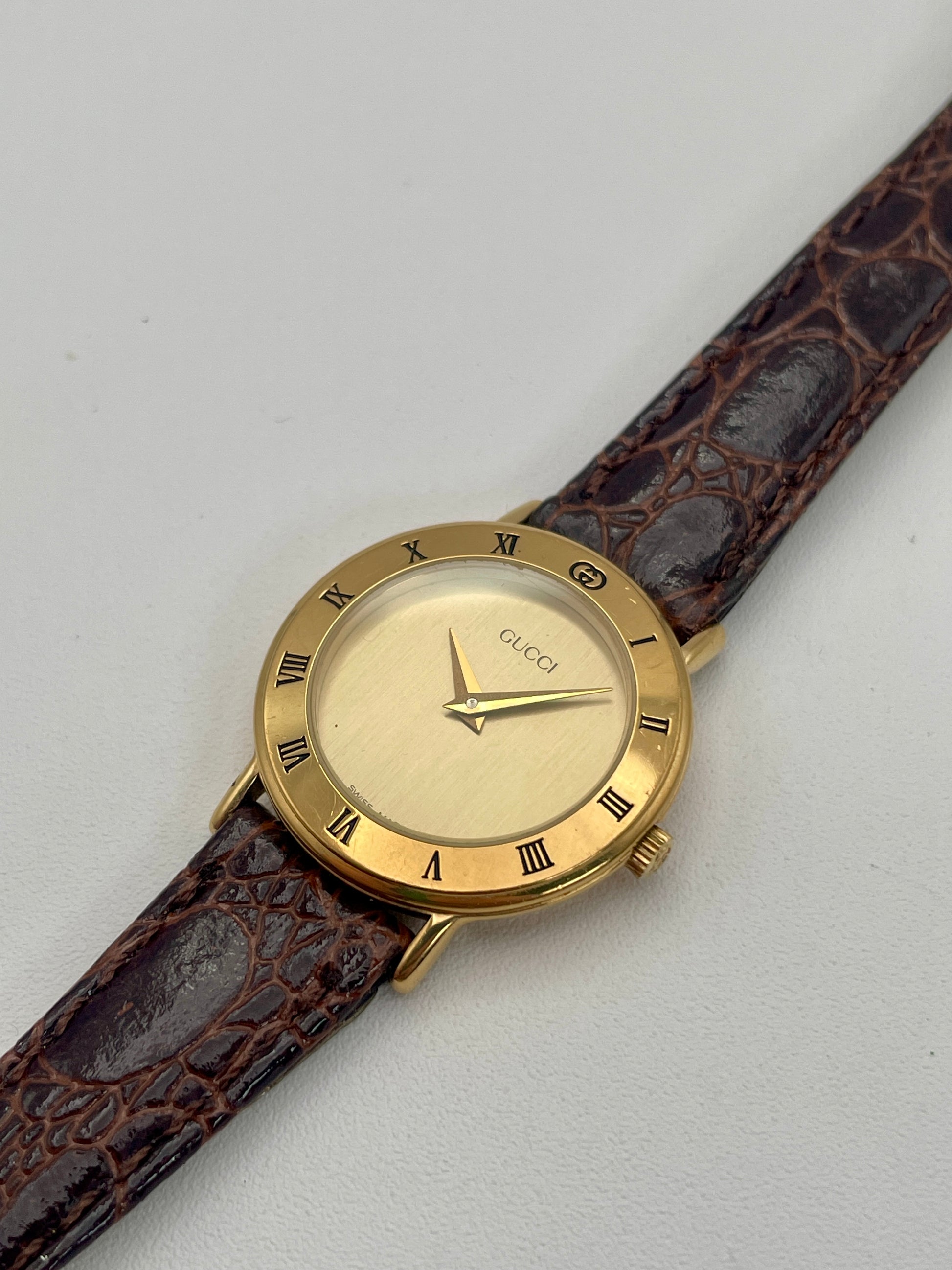 Diagonal view of the Gucci 3000.2.L watch featuring a gold-tone case, Roman numerals on the bezel, and a brown leather strap.