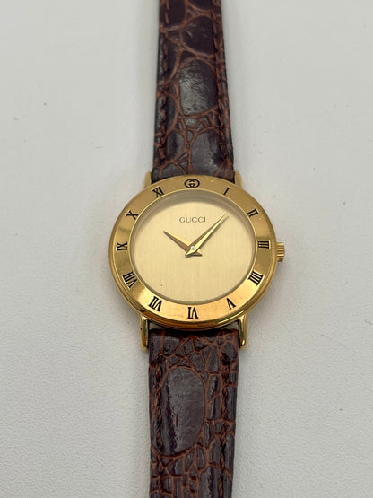 Front view of the Gucci 3000.2.L watch featuring a gold-tone case, Roman numerals on the bezel, and a brown leather strap.