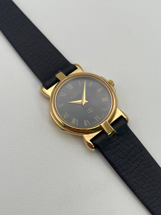 Gucci 3400F L Vintage Women's Watch in gold with a black leather strap, featuring a black dial with gold Roman numeral hour markers.