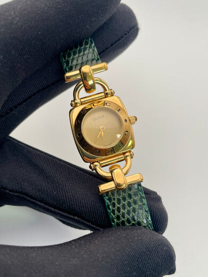 Close-up view of a Gucci 6300L ladies' watch, focusing on the  gold square case, gold dial, and green lizard skin strap.