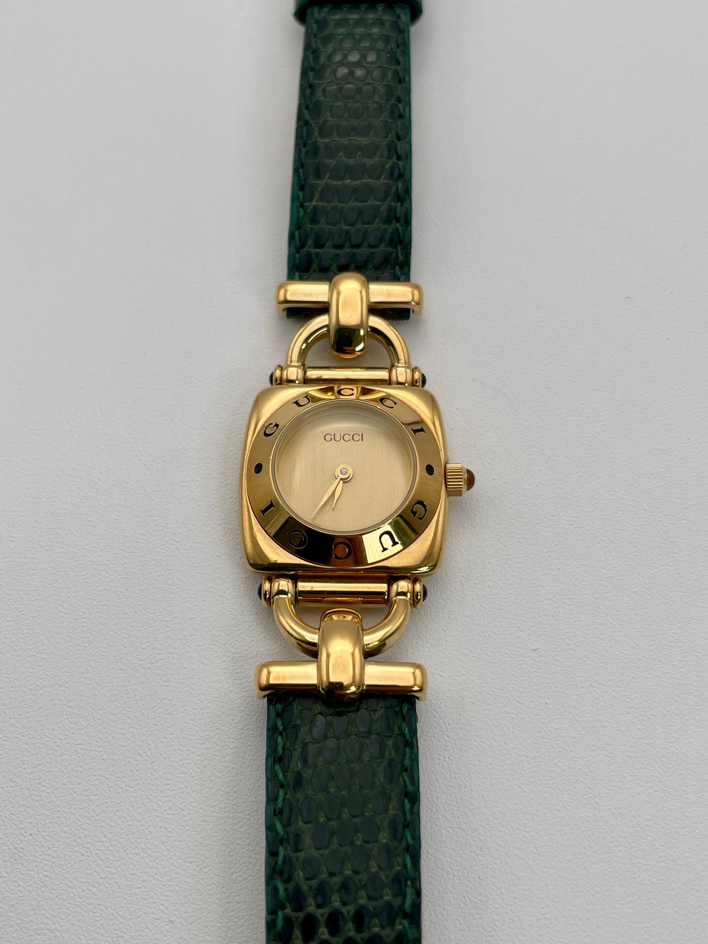 Front view of a Gucci 6300L ladies' watch featuring a  gold square case with rounded edges, gold dial, and green lizard skin strap.