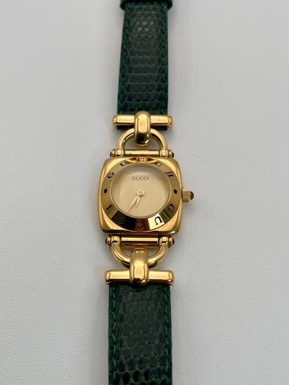 Front view of a Gucci 6300L ladies' watch featuring a  gold square case with rounded edges, gold dial, and green lizard skin strap.