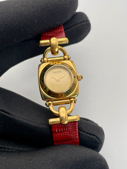 Close-up view of a Gucci 6300L ladies' watch, focusing on the gold-tone square case, gold dial, and red leather strap.