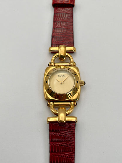 Front view of a Gucci 6300L ladies' watch featuring a gold-tone square case with rounded edges, gold dial, and red leather strap.