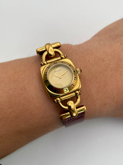 Gucci 6300L ladies' watch with a gold-tone square case, gold dial, and red leather strap, elegantly worn on the wrist, highlighting its refined design and fit.