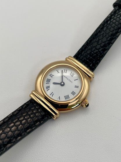 Diagonal view of Hamilton 8208 vintage gold watch with a round gold case, white Roman numeral dial, and black leather strap.