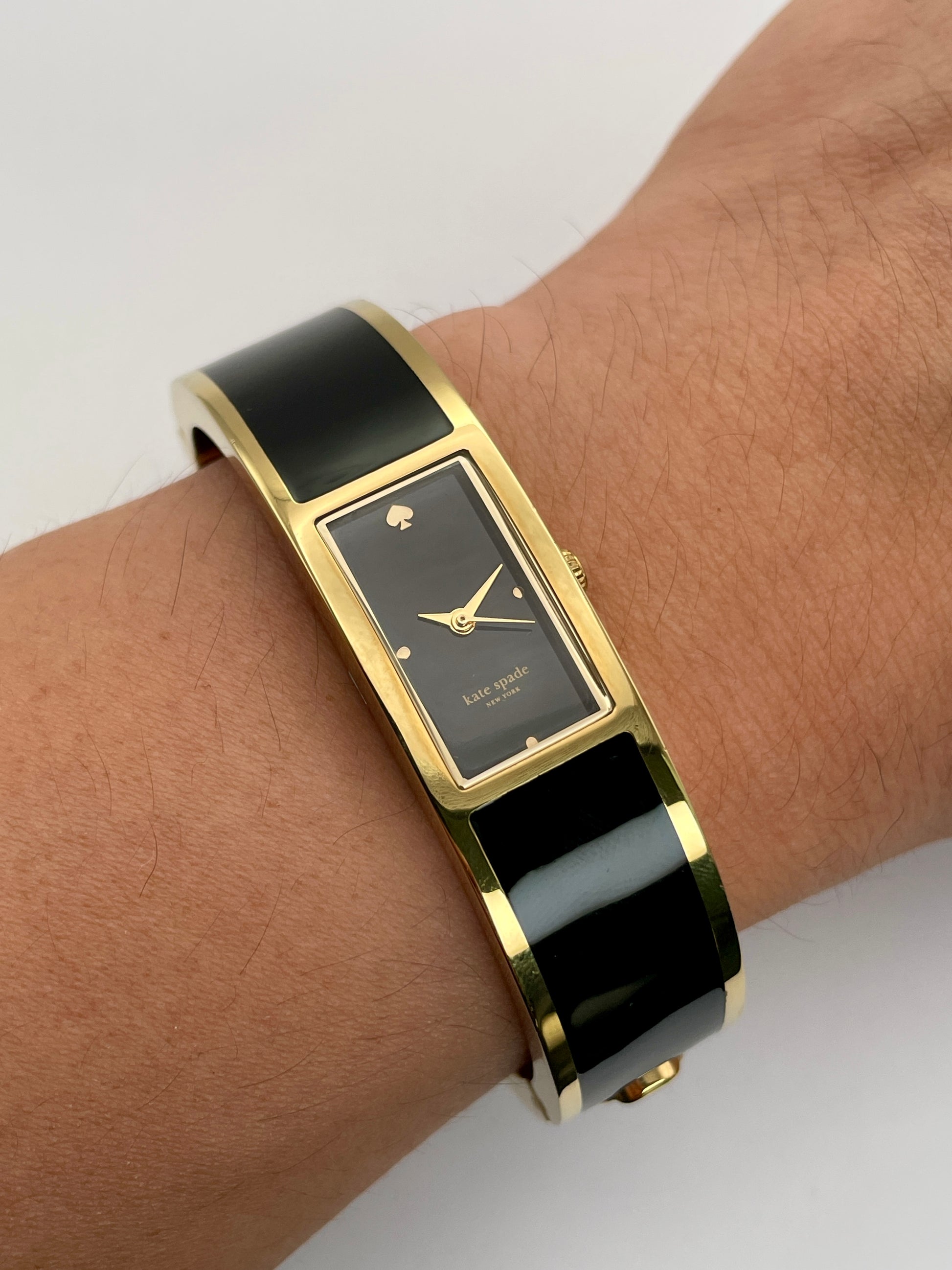 Wrist show view of * Kate Spade Bangle watch worn on wrist, featuring a gold-tone round case, black rectangular dial, and black enamel body.