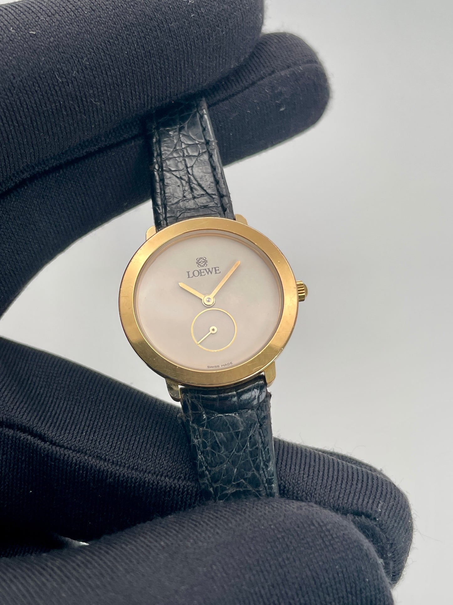 Gold watch with a black strap held by a black gloved hand on a light gray background. Another close-up emphasizing the gold hands and minimalist dial design.
