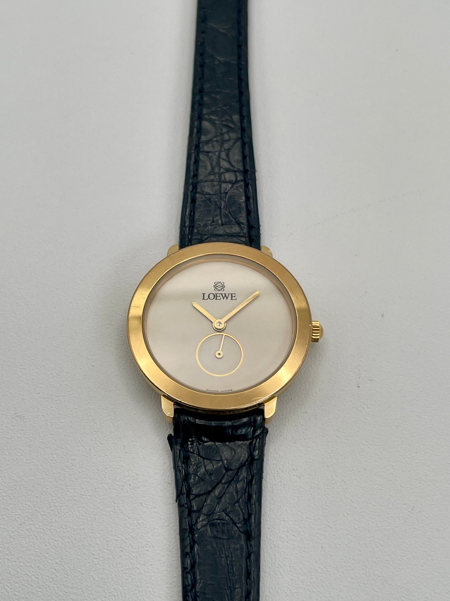 Gold watch with black leather strap on a gray background. Front view of Loewe Calatrava Vintage Women's Watch in gold case with black leather strap showing white dial and small seconds subdial.