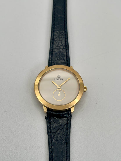 Gold watch with black leather strap on a gray background. Front view of Loewe Calatrava Vintage Women's Watch in gold case with black leather strap showing white dial and small seconds subdial.