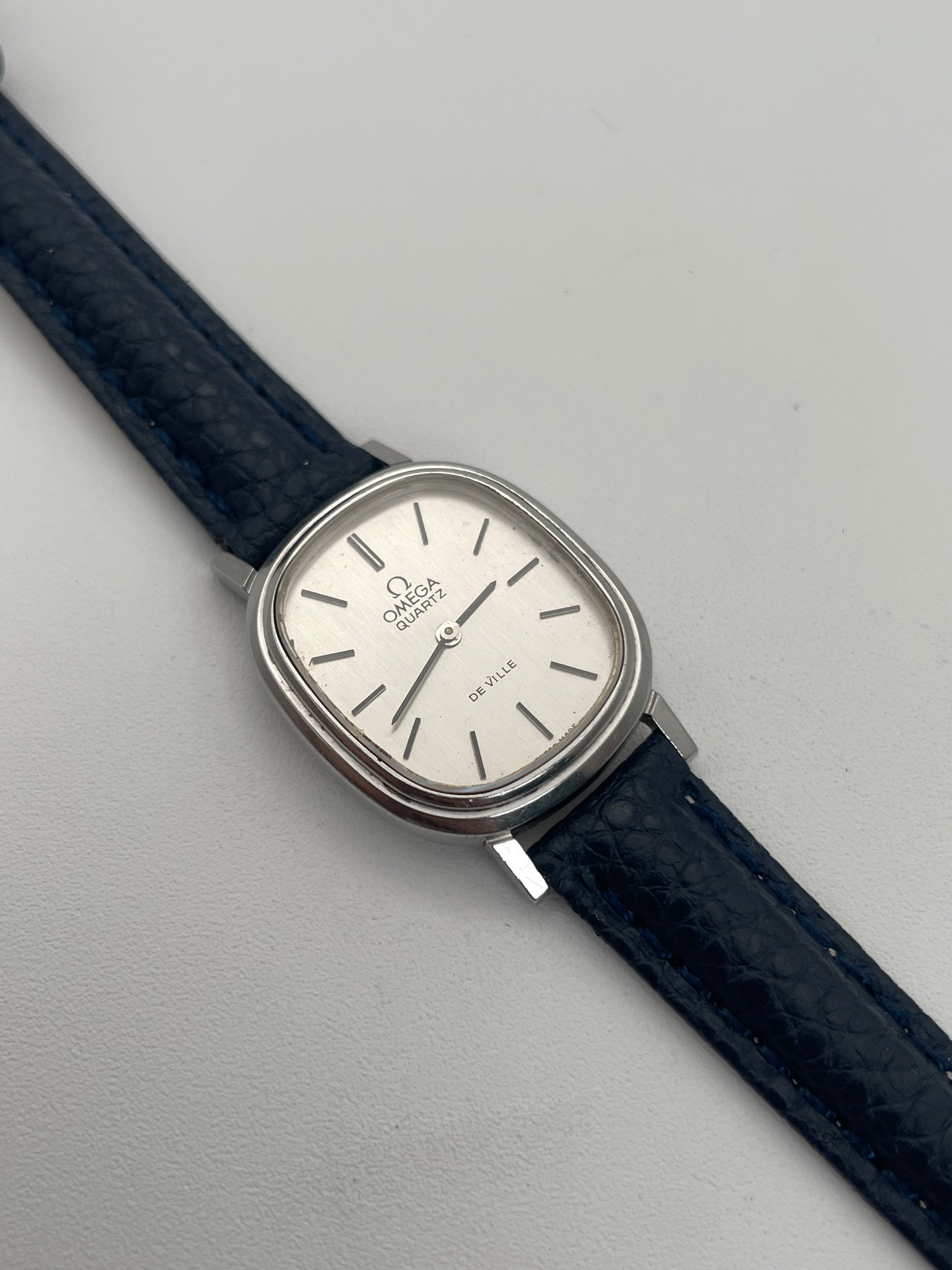 Omega 591.0016 vintage quartz watch with an oval-shaped silver case, rectangular silver dial, slim black indices, and blue leather strap.