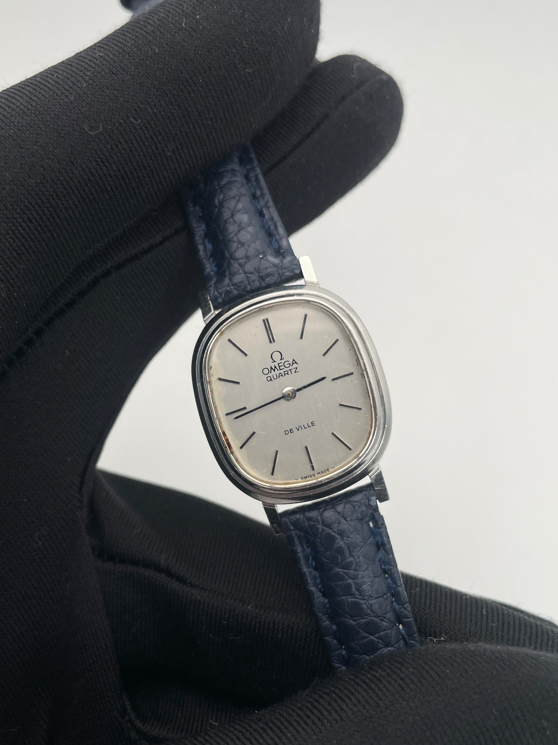 Close-up view of Omega 591.0016 vintage quartz watch with an oval-shaped silver case, rectangular silver dial, slim black indices, and blue leather strap.