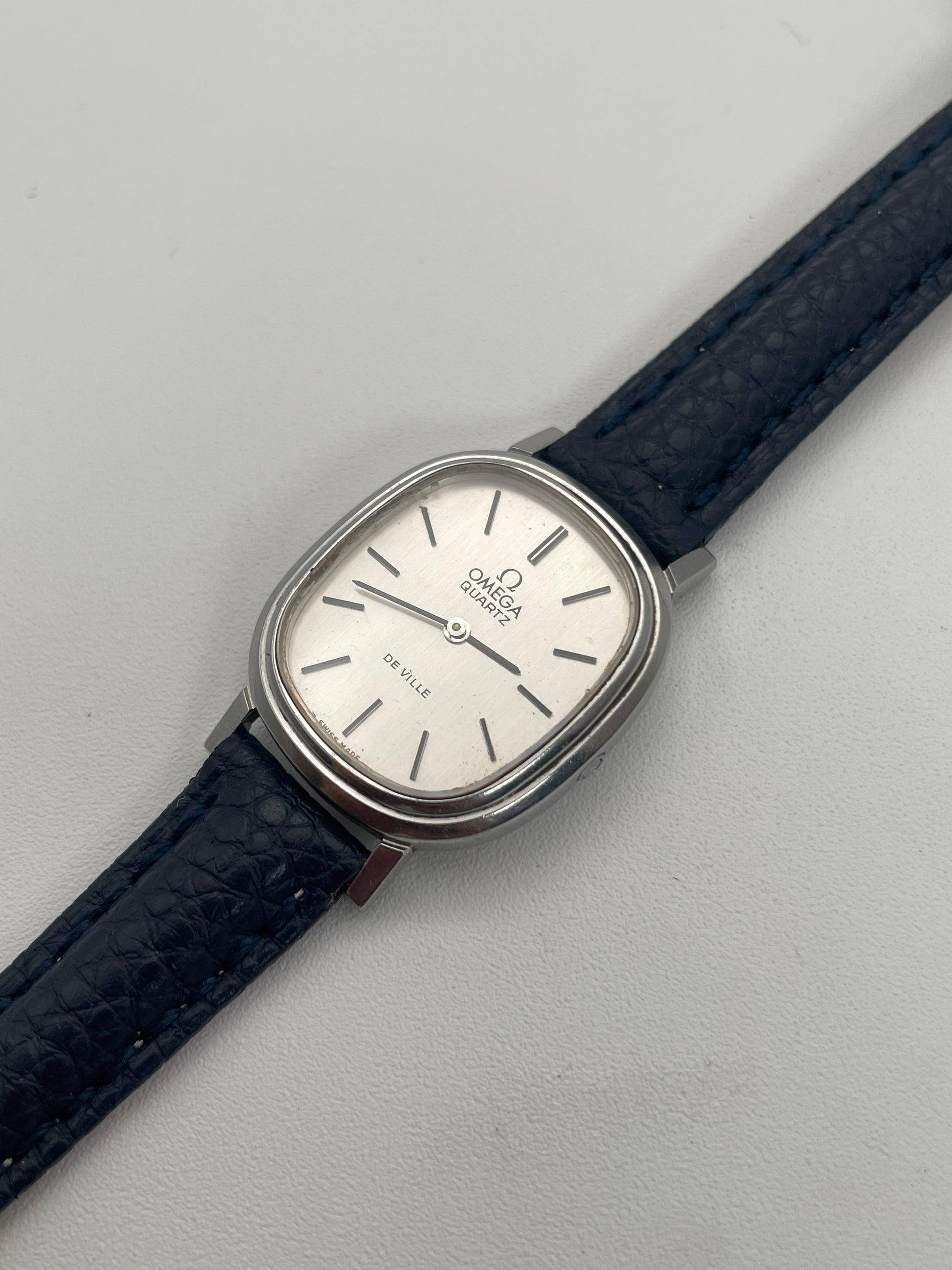 Diagonal view of Omega 591.0016 vintage quartz watch with an oval-shaped silver case, rectangular silver dial, slim black indices, and blue leather strap.