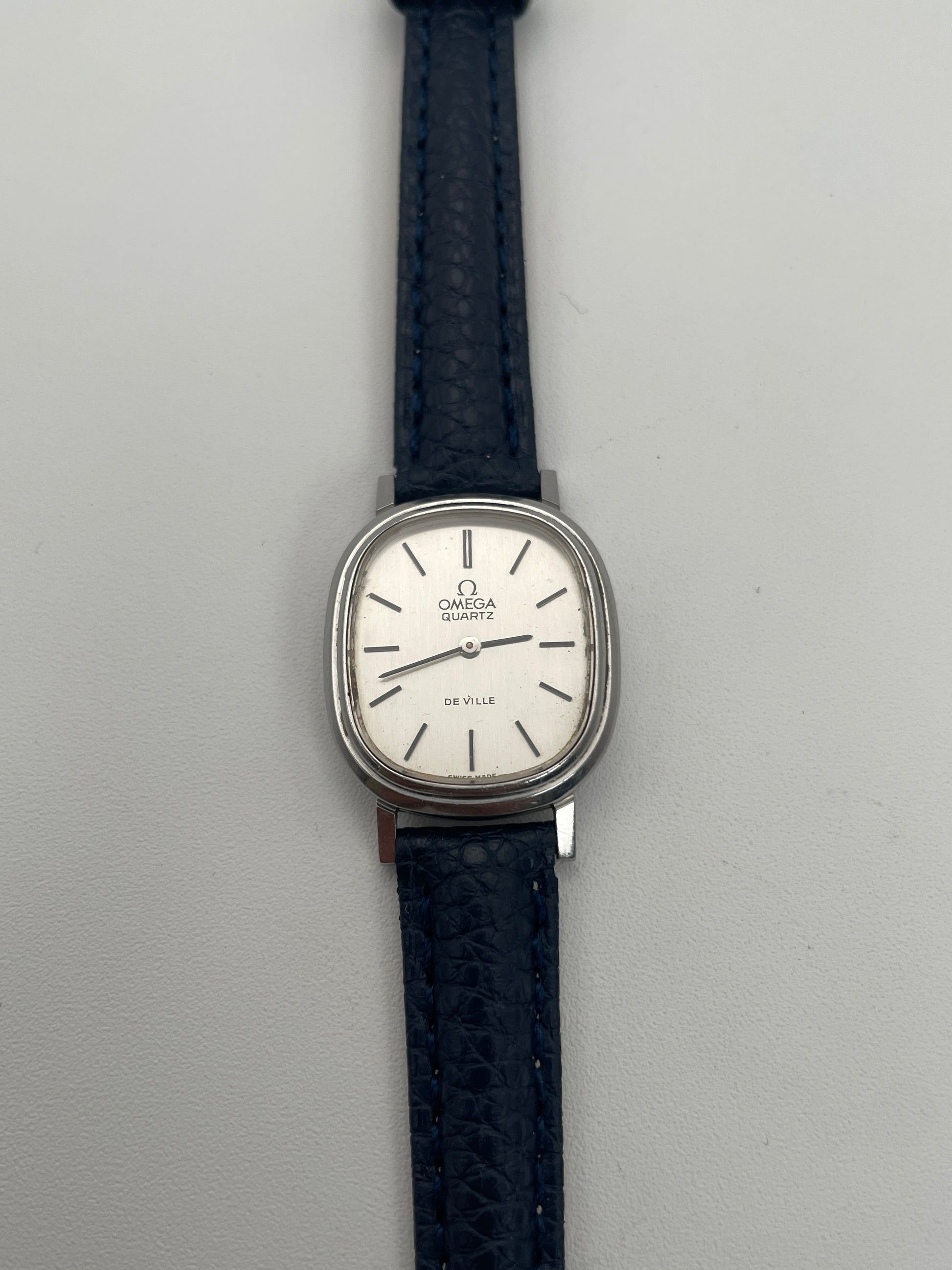 Front view of Omega 591.0016 vintage quartz watch with an oval-shaped silver case, rectangular silver dial, slim black indices, and blue leather strap.