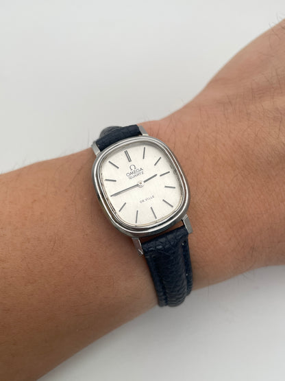 Wrist shot of Omega 591.0016 vintage quartz watch with an oval-shaped silver case, rectangular silver dial, slim black indices, and blue leather strap.