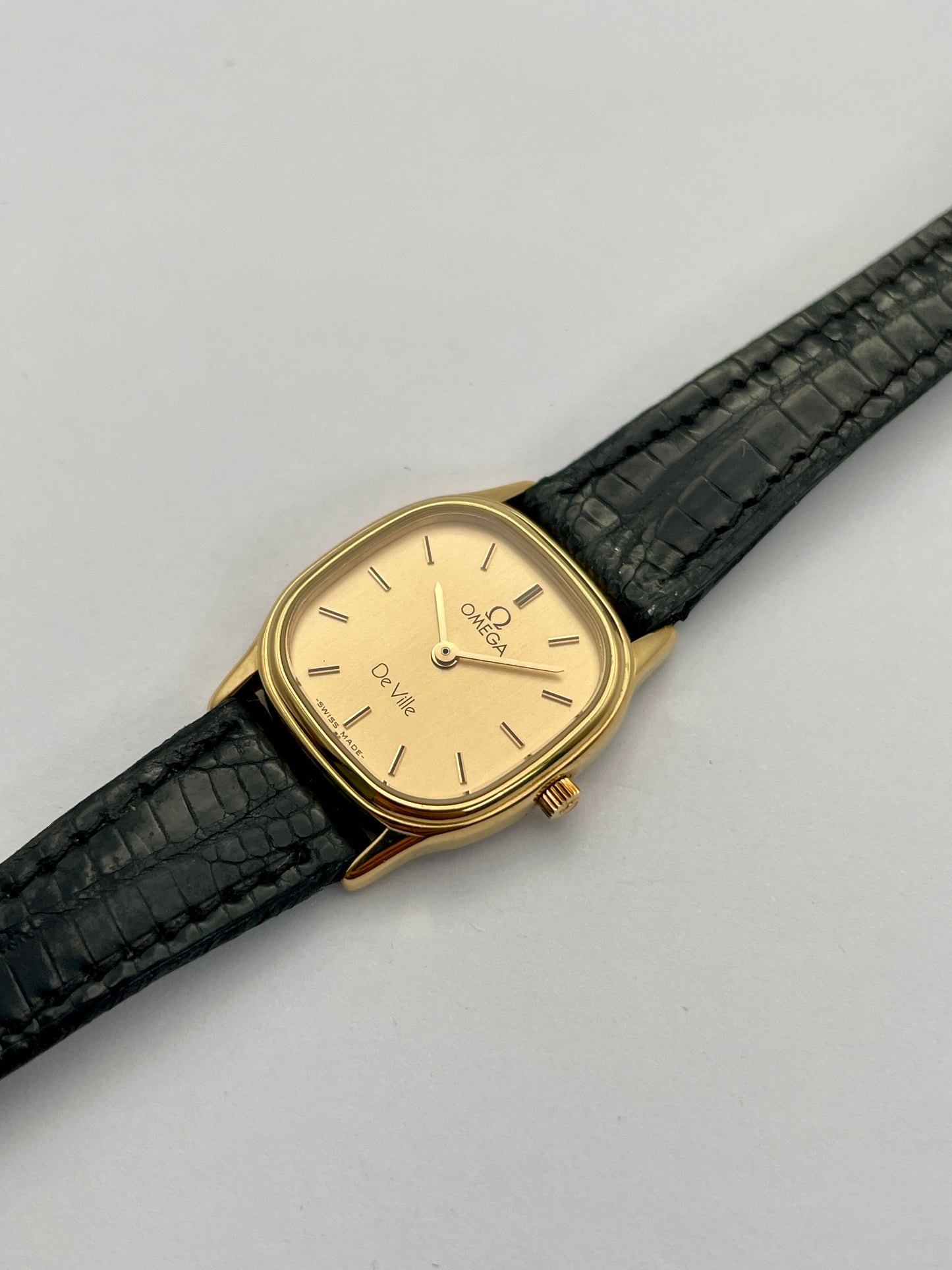 Diagonal view of Omega De Ville vintage gold watch with a square case, champagne dial, and black leather strap.