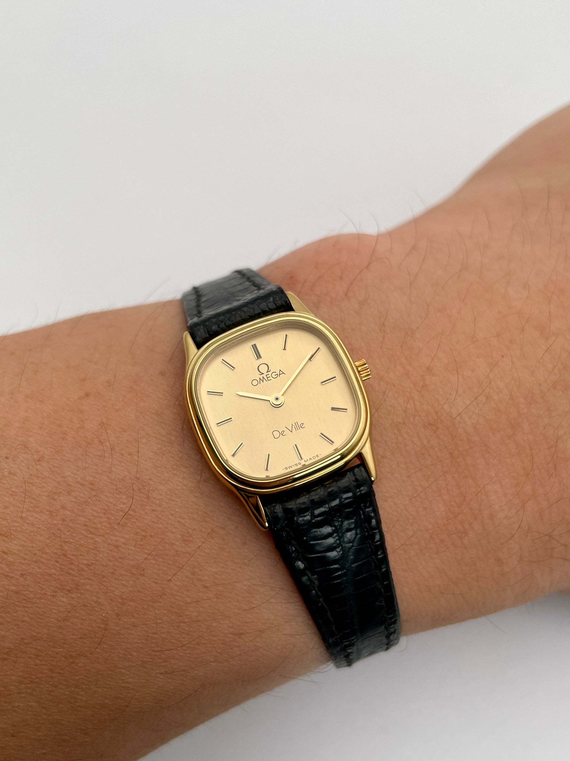 Wrist shot of Omega De Ville vintage gold watch with a square gold case, champagne dial, and black leather strap.