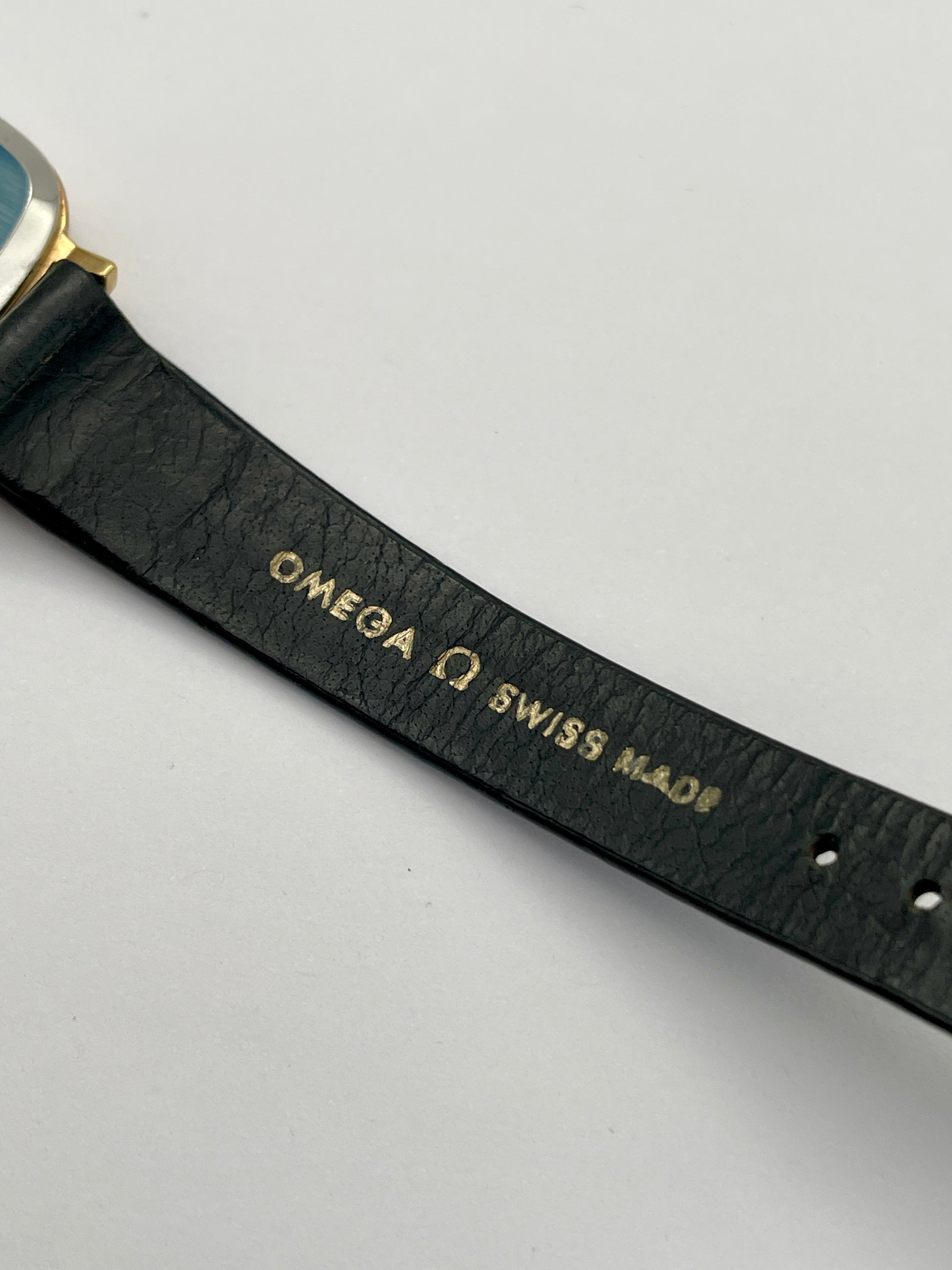 Close-up view of Omega De Ville TV Dial strap showcasing original Omega strap and branding, watch with a gold-tone square case and black leather strap.