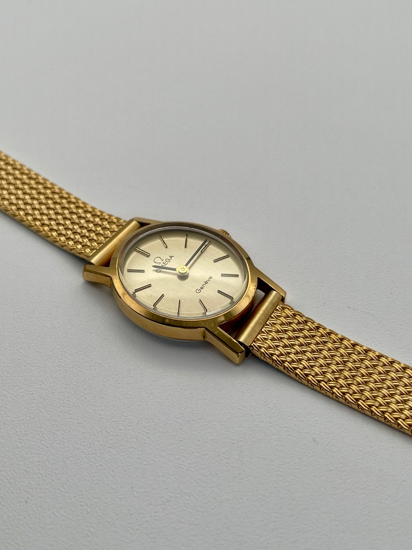 Omega Genève oval ladies watch in gold with a mesh bracelet, flatlay view.
