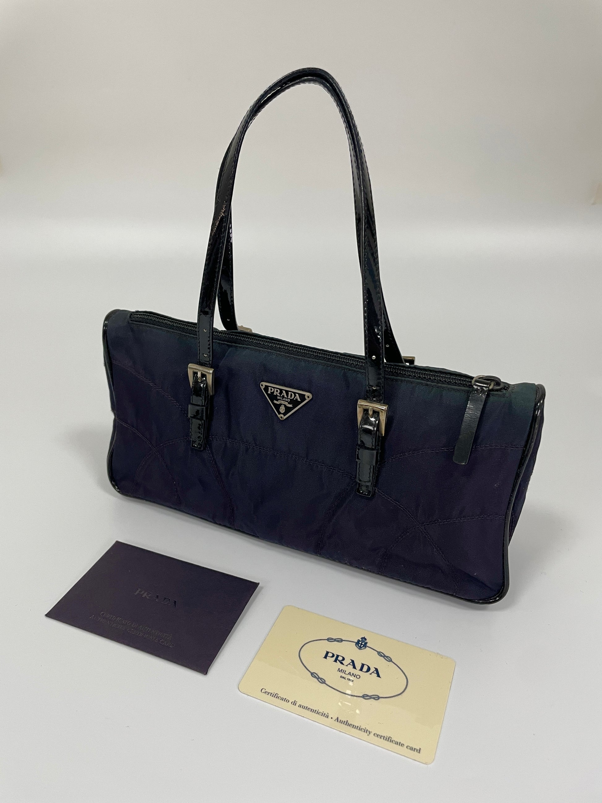 Angled view of a Prada Tessuto Spazzolato bag in indigo prune nylon with leather handles and two buckle details, displayed with authenticity cards.