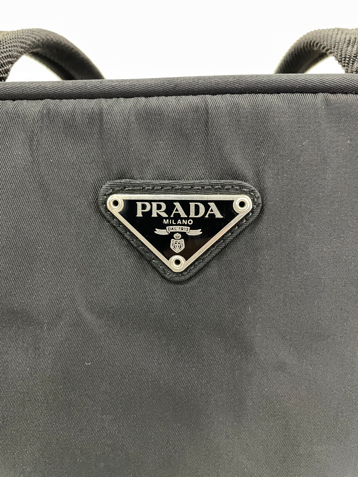 Close-up view of the triangular Prada logo plate on the Prada Tessuto Tote Bag in black nylon.