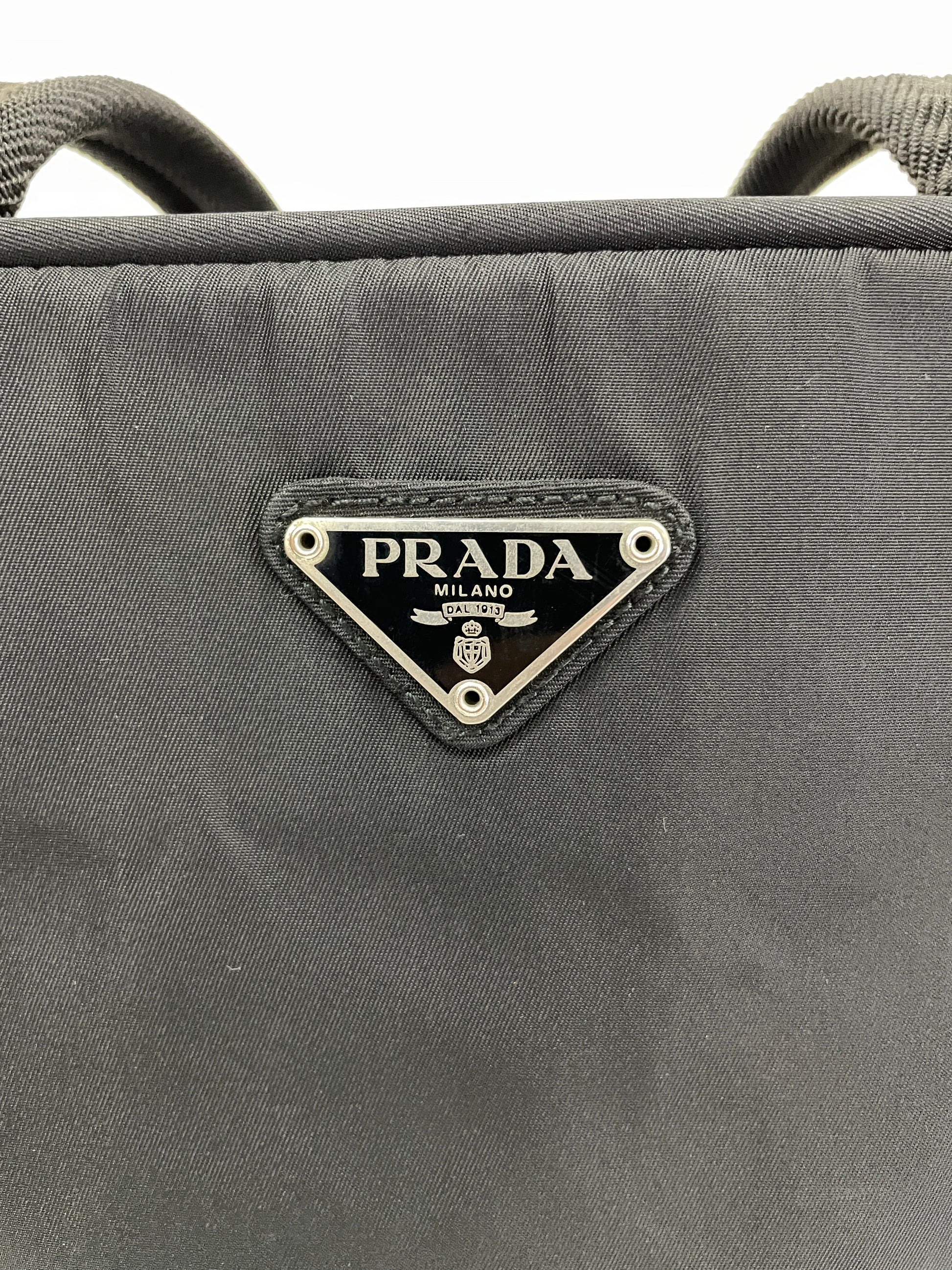 Close-up view of the triangular Prada logo plate on the Prada Tessuto Tote Bag in black nylon.