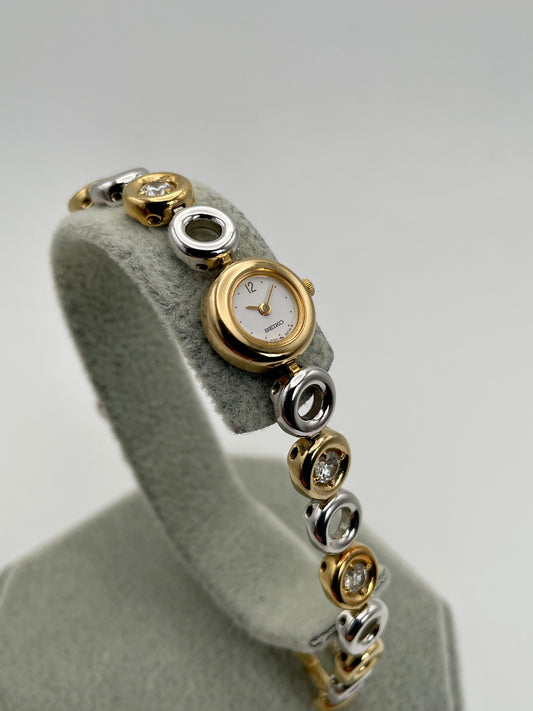 Seiko 1E20-0020 vintage women’s watch in steel and gold with crystal-accented bracelet, displayed on stand
