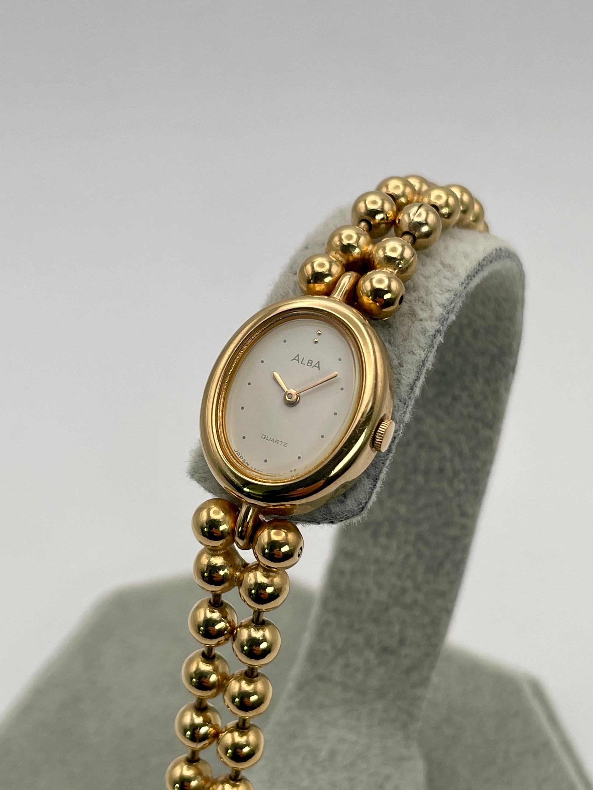 Gold watch with a beaded strap on a gray stand against a white background. Diagonal right view of Seiko Alba Y588-5180 vintage watch with reflective gold finish and clean dial.